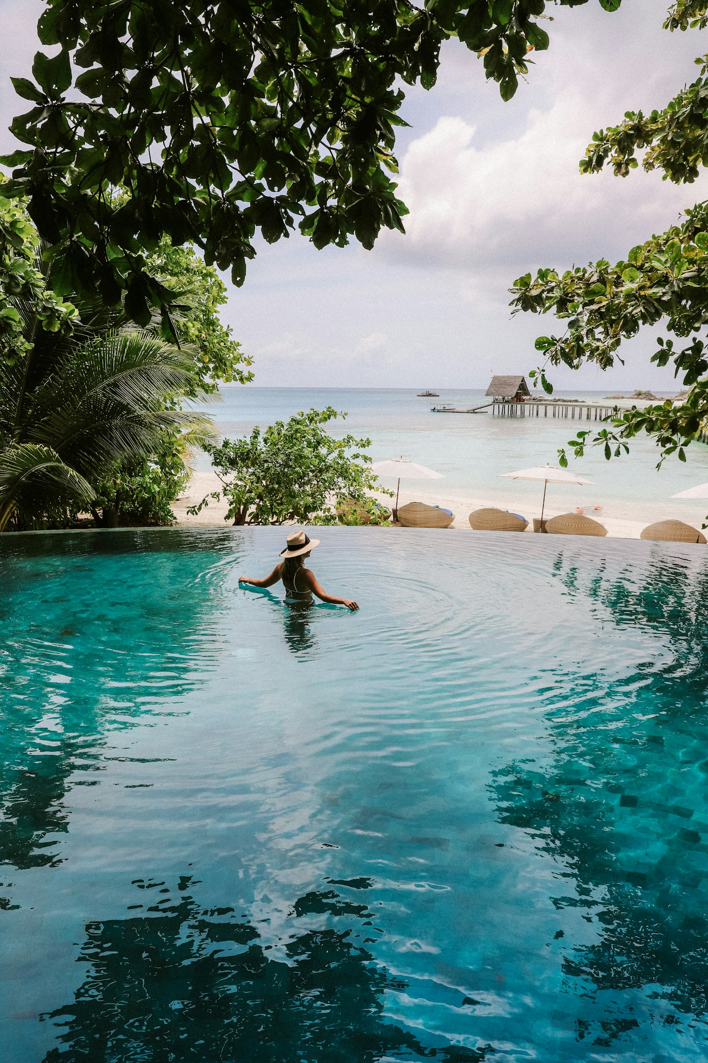 A traveler in a pool overlooking a palm-framed beach and turquoise water